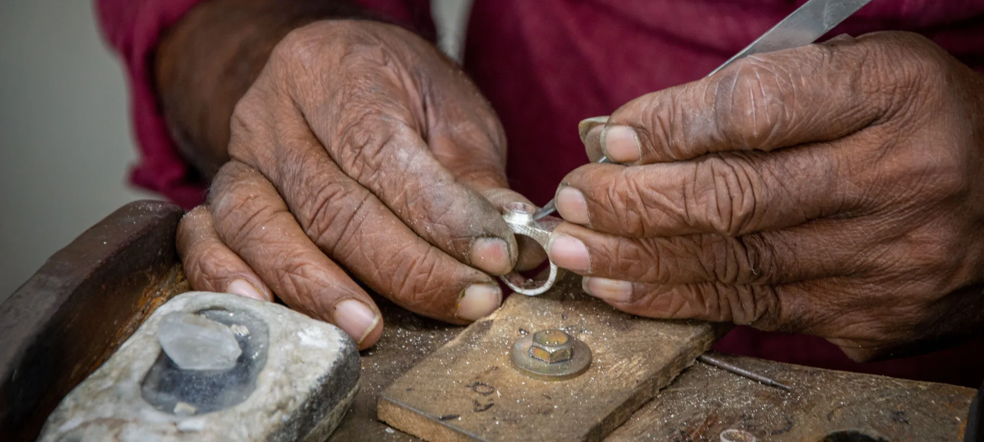 Le mani segnate dal tempo di un artigiano che tengono un piccolo anello d'argento e uno strumento appuntito su un blocco da banco in legno